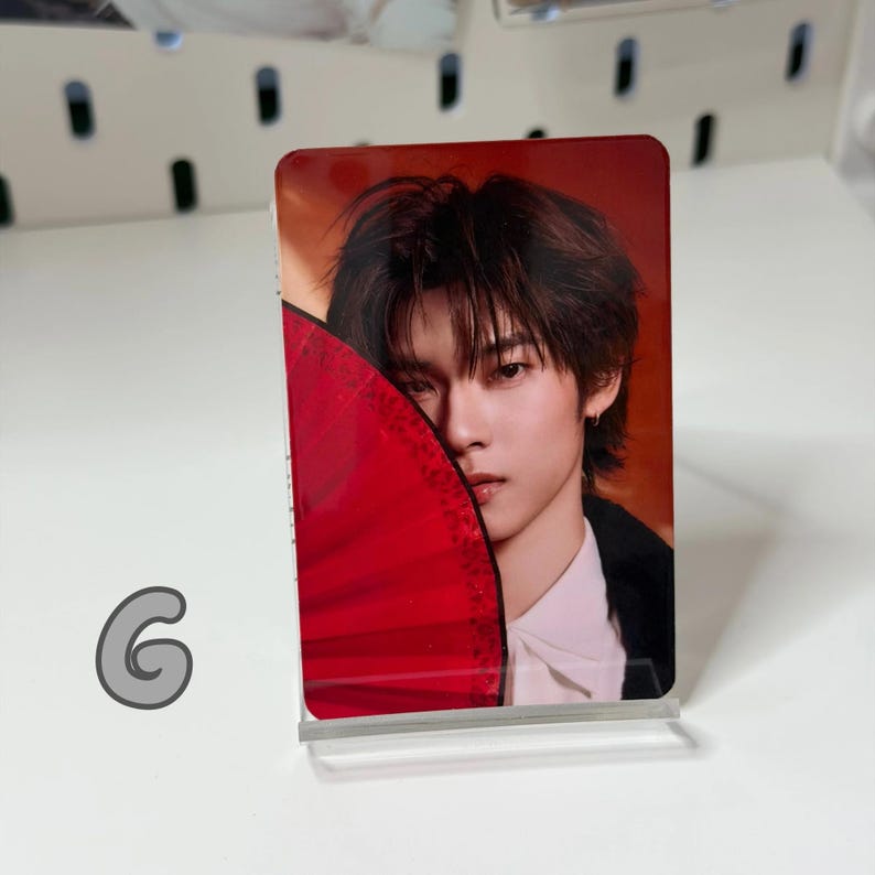 May include: A rectangular card featuring a person with dark hair and a red fan. The person is wearing a white shirt and black jacket. The card is displayed on a clear acrylic stand.