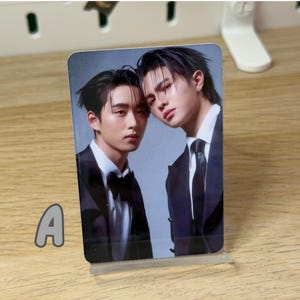 May include: A photograph of two people in formal attire, likely a promotional image. The subjects are wearing black suits with white shirts, one with a bow tie and the other with a necktie. The image is displayed in a clear acrylic stand.