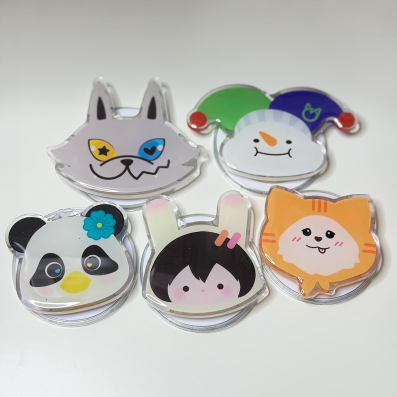 May include: A collection of five clear acrylic phone grips featuring cartoon animal and character designs. The designs include a wolf, a snowman, a panda, a girl with bunny ears, and an orange cat.