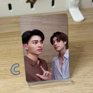 May include: A rectangular photo card featuring two young men. One is wearing a brown shirt and the other a light blue button-down shirt. The card is propped up on a clear plastic stand, with the letter "C" in a light gray color in the lower left corner.