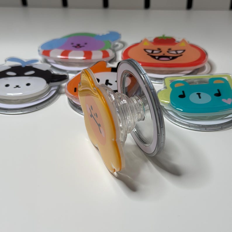 May include: A collection of colorful phone grips with various cartoon character designs. The grips feature a clear, expandable base and a circular, adhesive backing. Designs include a bear, cat, and other cute characters in shades of orange, blue, and black.