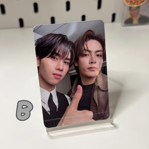 May include: A clear acrylic photo stand holding a photo of two young men. One man is wearing a black vest and tie, the other a brown jacket. The photo is a selfie with a thumbs-up gesture.