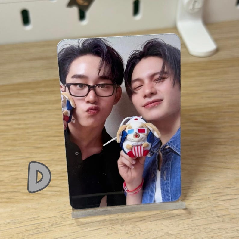 May include: A clear acrylic photo stand displaying a photograph of two young men. One man wears glasses and makes a kissing face. The other man holds a plush dog wearing 3D glasses. The photo is on a wooden surface.