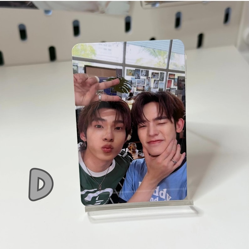 May include: A photo card featuring two young men posing for a selfie. One man is wearing a green shirt, the other a blue shirt. The photo card is displayed on a clear acrylic stand.