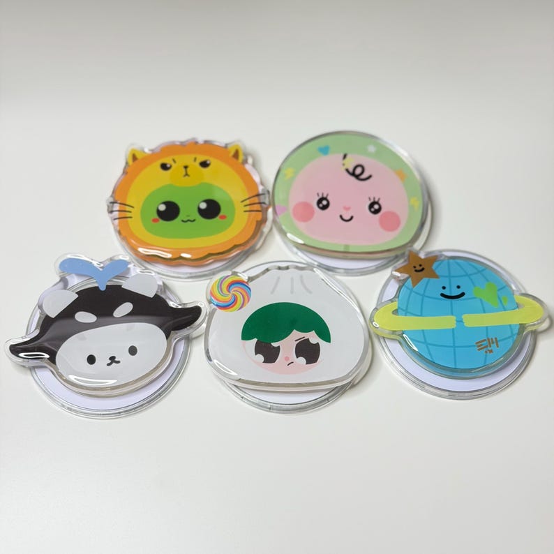 May include: Five clear acrylic pins with cartoon character designs. The pins feature a cat, a peach, a Shiba Inu, a dumpling, and a planet with a yellow ring. Each pin has a white backing and colorful illustrations.