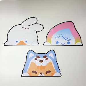 Op de afbeelding: Drie stickers in cartoonstijl met dierontwerpen. Eén is een wit konijn, een ander een regenboogkleurig personage en de laatste een blauwe en oranje kat. Elke sticker heeft een zwarte omtrek en een schattig, minimalistisch ontwerp.