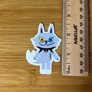 Op de afbeelding: Een sticker met een cartoonwolf met een lichtblauw lichaam, een zwarte kraag en een witte staart. De wolf heeft een geel ster-oog en een blauw hart-oog. De sticker is ongeveer 5 cm hoog.