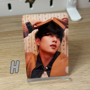 May include: A clear acrylic photo stand holding a photo of a person with a book on their head. The person is wearing a brown jacket over a denim jacket. The letter "H" is visible in the lower left corner.