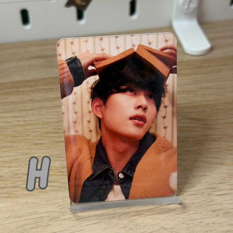 May include: A clear acrylic photo stand holding a photo of a person with a book on their head. The person is wearing a brown jacket over a denim jacket. The letter "H" is visible in the lower left corner.