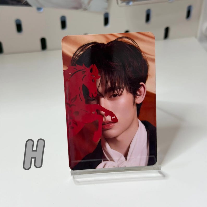 May include: A rectangular photo card featuring a person with dark hair and a red horse silhouette overlay. The card is displayed on a clear acrylic stand. The letter "H" is visible in the lower left corner.