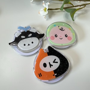 May include: Three colorful phone grips with cartoon designs. One features a black and white cow, another a baby with a green background, and the third a black and orange cat-like character. Each grip has a clear, circular base.