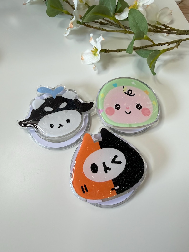 May include: Three colorful phone grips with cartoon designs. One features a black and white cow, another a baby with a green background, and the third a black and orange cat-like character. Each grip has a clear, circular base.