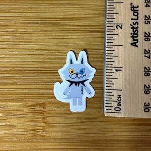 Op de afbeelding: Een sticker met een cartoonwolf met een lichtblauw lichaam, witte accenten en een zwarte kraag. De wolf heeft &eacute;&eacute;n geel en &eacute;&eacute;n blauw oog. De sticker is ongeveer 2,5 cm hoog.