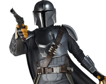 Könnte beinhalten: Eine detaillierte Actionfigur eines mandalorianischen Kriegers aus dem Star Wars-Universum. Die Figur trägt eine silberne und schwarze Rüstung mit einem braunen Ledergürtel und einem Umhang. Die Figur hält eine Blasterpistole in der rechten Hand und steht auf einem Sandsockel.