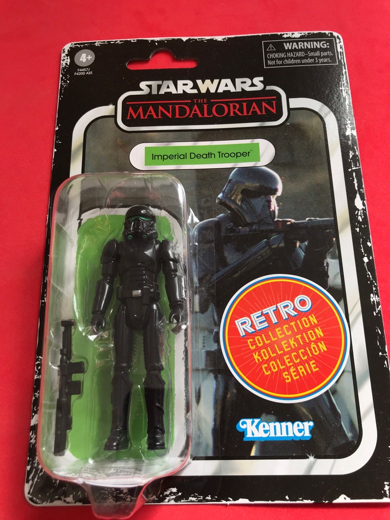 May include: A Star Wars The Mandalorian Imperial Death Trooper action figure in a retro-style package. The black figure is in a clear plastic bubble, with a black rifle. The packaging includes the text "RETRO COLLECTION" and the Kenner logo.