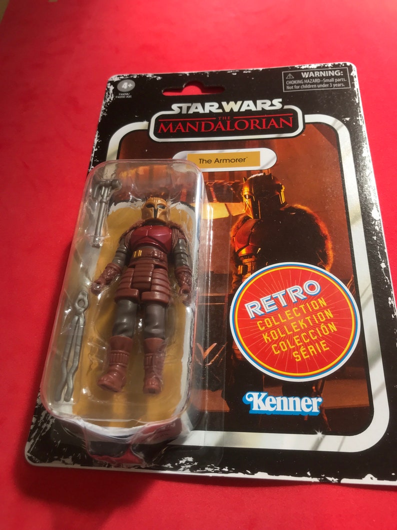 Pu&ograve; includere: Una action figure di Star Wars The Mandalorian The Armorer in una confezione retr&ograve; Kenner. La figura indossa un'armatura rossa e marrone con un elmetto e tiene un'arma. La confezione ha un logo Kenner retr&ograve; e il testo "Collezione Retro" in pi&ugrave; lingue.