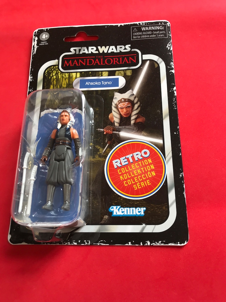 Puede incluir: Una figura de acci&oacute;n de Star Wars: The Mandalorian de Ahsoka Tano en un paquete de estilo retro. La figura est&aacute; en una caja de pl&aacute;stico transparente, con una tarjeta en blanco y negro con el logotipo de Star Wars, el nombre de Ahsoka Tano y una pegatina de "Retro Collection".