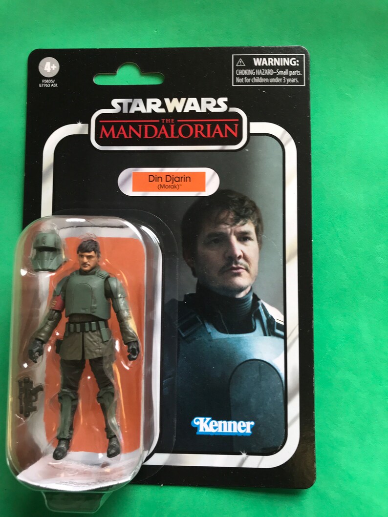 May include: A Star Wars The Mandalorian action figure of Din Djarin (The Mandalorian) in a green and brown outfit with a helmet. The figure is packaged in a retro Kenner style box with a photo of Pedro Pascal as Din Djarin on the front.