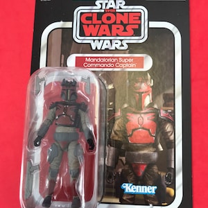 May include: A Star Wars The Clone Wars action figure of a Mandalorian Super Commando Captain. The figure is in a plastic blister pack with a red, white, and blue Kenner logo. The figure is wearing red and grey armour with a helmet. The figure is holding a blaster rifle.