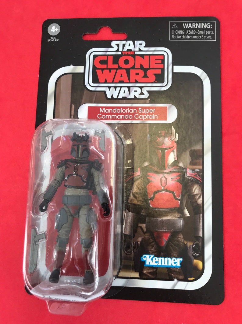May include: A Star Wars The Clone Wars action figure of a Mandalorian Super Commando Captain. The figure is in a plastic blister pack with a red, white, and blue Kenner logo. The figure is wearing a red and grey armor with a helmet. The figure is holding a blaster rifle.