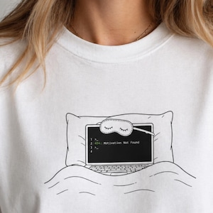 May include: A white t-shirt featuring a black line art graphic of a laptop tucked into bed with a pillow and blanket. The laptop screen displays the text 404: Motivation Not Found and is wearing a sleep mask over the top edge of the screen.