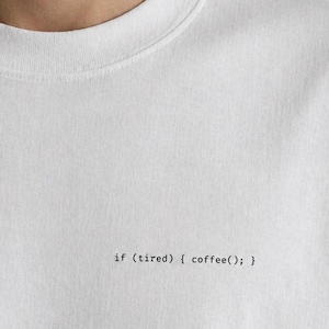 Op de afbeelding: Een wit t-shirt met korte mouwen en kleine zwarte tekst op de borst die luidt: if (tired) { coffee(); } Het shirt heeft een ronde hals en een eenvoudig ontwerp met de programmeercode gedrukt in een lettertype dat lijkt op een typemachine.
