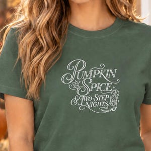 May include: A dark green short-sleeved t-shirt featuring white text that reads Pumpkin Spice & Two-Step Nights, accompanied by a small leaf graphic and a line-art illustration of a cowboy boot with decorative stars.