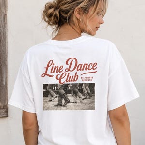 Può includere: Una t-shirt bianca a maniche corte con testo rosso che recita Line Dance Club e No Partner Required sopra una fotografia in bianco e nero di persone con stivali da cowboy che ballano su un pavimento in legno, abbinata a pantaloncini di jeans.