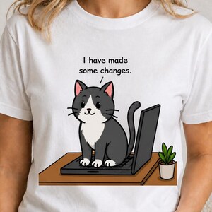 Pode incluir: Uma t-shirt branca de manga curta com um gráfico de um gato cinzento e branco sentado num portátil aberto numa secretária castanha. O gato tem um balão de fala acima da cabeça com o texto, I have made some changes. Um pequeno vaso com uma planta verde está ao lado.