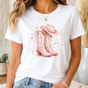 Könnte beinhalten: Ein weißes Kurzarm-T-Shirt mit einem Grafikdruck von zwei hellrosa Cowboystiefeln und einem passenden rosa Cowboyhut. Das Design ist mit kleinen rosa Herzen, Musiknoten und geschwungenen dekorativen Linien im Aquarellstil verziert.