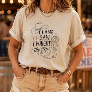 May include: A beige short-sleeved t-shirt featuring a black graphic print of a cowboy hat and a western boot with the text "I CAME, I SAW, I FORGOT the steps" surrounded by small star icons. The shirt has a crew neckline and rolled sleeves.
