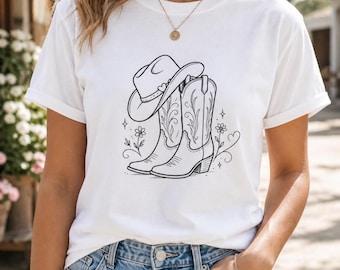 May include: A white short-sleeved t-shirt featuring a black line art graphic on the chest. The design depicts a cowboy hat resting on a pair of cowboy boots, surrounded by small floral accents, stars, and a heart shape, creating a simple illustration.