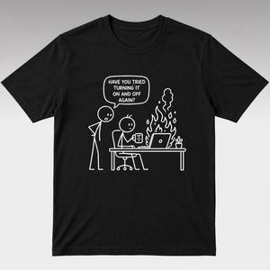 Könnte beinhalten: Schwarzes T-Shirt mit einer weißen Grafik von Strichmännchen. Einer fragt: „HABEN SIE VERSUCHT, ES AUS- UND WIEDER EINZUSCHALTEN?“ während der Computer eines anderen brennt. Eine Kaffeetasse und eine Pflanze stehen auf dem Schreibtisch.