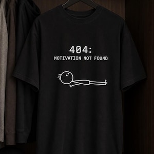 Könnte beinhalten: Ein schwarzes T-Shirt mit kurzen Ärmeln und dem weißen Text 404: MOTIVATION NOT FOUND. Unter dem Text befindet sich eine einfache weiße Strichzeichnung einer Strichmännchen-Figur, die flach auf dem Bauch liegt und horizontal ausgerichtet ist.