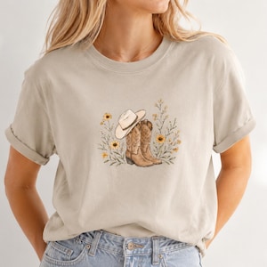 May include: A beige short-sleeve t-shirt featuring a central graphic illustration of a light-colored cowboy hat resting on a pair of brown leather cowboy boots, surrounded by a cluster of yellow sunflowers and green leafy stems.