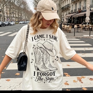 Pode incluir: Uma t-shirt em tom creme com uma estampa gráfica preta de um chapéu de cowboy e duas botas de cowboy com detalhes florais. O texto diz I CAME, I SAW, I FORGOT The Steps. A peça é combinada com um boné bege e uma bolsa preta de ombro.