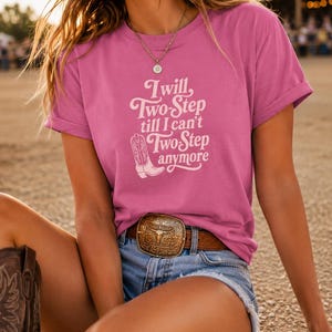 Puede incluir: Una camiseta rosa de manga corta con el texto blanco 'I will, Two-Step till I can't Two-Step anymore' y un gráfico de una bota vaquera. La camiseta se combina con pantalones cortos de mezclilla azul y un cinturón de cuero marrón.