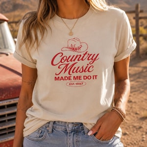 May include: A cream-colored short-sleeve t-shirt featuring a red graphic print of a cowboy hat above the text "Country Music MADE ME DO IT" and "EST. 1987" enclosed in an oval. The shirt has a crew neckline and is worn with blue denim shorts.