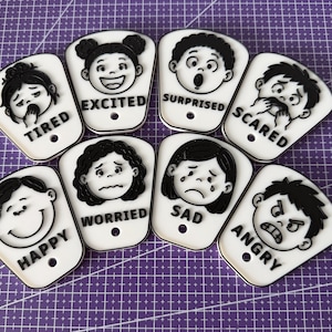 May include: A set of eight white acrylic tags with black raised illustrations of faces and text labels. Each tag features a specific emotion: Tired, Excited, Surprised, Scared, Happy, Worried, Sad, and Angry. Each piece has a small circular hole.