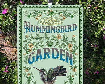 Puede incluir: Un letrero metálico rectangular con las palabras "Hummingbird Garden" en azul y verde, adornado con ilustraciones florales y de colibríes. El letrero tiene un fondo verde claro y un borde decorativo. Montado sobre una estaca de metal.