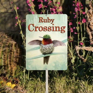 May include: A green, red, and black hummingbird with a long beak is on a sign that says "Ruby Crossing". The sign is on a metal pole and is surrounded by green plants.