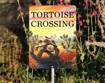 May include: A yellow and orange sign with the text "TORTOISE CROSSING" in black. The sign features a painted image of a tortoise walking across a desert landscape with a hummingbird flying above.