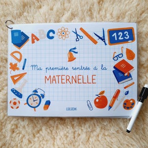May include: A white board with a blue and orange border. The board has a grid pattern and is decorated with school-themed illustrations, including letters, numbers, a bell, a ruler, a pencil, and a book. The text "Ma première rentrée à la MATERNELLE" is written in blue cursive on the board. The text "LULUDIK" is written in blue at the bottom of the board.
