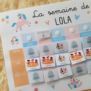 May include: A colorful weekly schedule chart for a child named Lola. The chart features a unicorn illustration, a rainbow, and various icons representing activities like school, daycare, and meals. The days of the week are labeled in French.