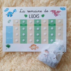 May include: A white weekly chart with green and beige sections. The chart is labeled "La semaine de LUCAS" and has columns for each day of the week. There are spaces for stickers to be placed in each column. The chart is decorated with a blue dinosaur and a pink dinosaur.