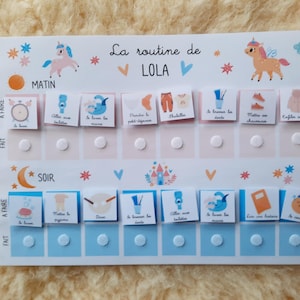 May include: A white board with a blue and pink color scheme. The board has a daily routine chart for a child named Lola. The chart includes activities like brushing teeth, getting dressed, and having breakfast. The board has a unicorn illustration at the top.