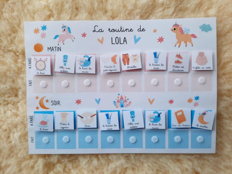 May include: A white board with a blue and pink color scheme. The board has a daily routine chart for a child named Lola. The chart includes activities like brushing teeth, getting dressed, and having breakfast. The board has a unicorn illustration at the top.