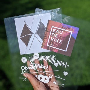 May include: A clear acrylic piece with white text and music-themed graphics, including play buttons and a sound wave. The text includes "Obsession", "EXO", and "Scenarist". Additional cards feature band logos and the text "EAU DE VIXX".