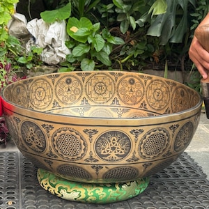 Puede incluir: Un cuenco tibetano grande y ornamentado con intrincados patrones grabados en negro y dorado. El cuenco descansa sobre un cojín verde y dorado. También hay un mazo de madera y un mazo rojo.