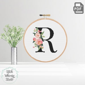 Könnte beinhalten: Ein Kreuzstichmuster des Buchstabens R mit rosa und pfirsichfarbenen Blumen. Das Muster befindet sich in einem hölzernen Stickrahmen.